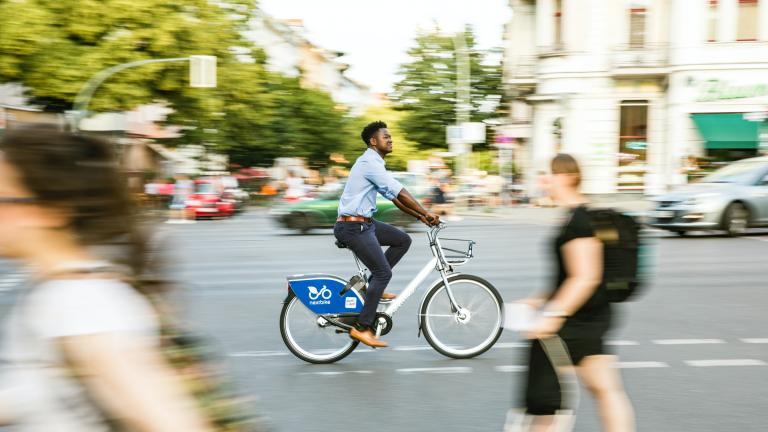 An image of a street scene with pedestrians and a cyclist. Trees are seen in the background.